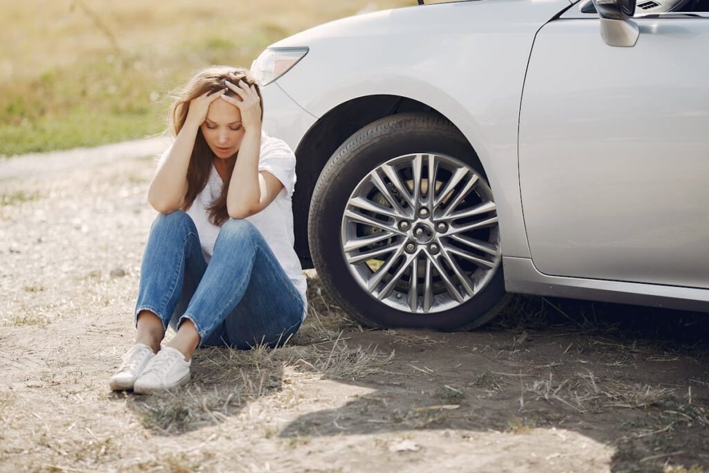 Worried young woman sitting near broken automobile at roadside in countryside